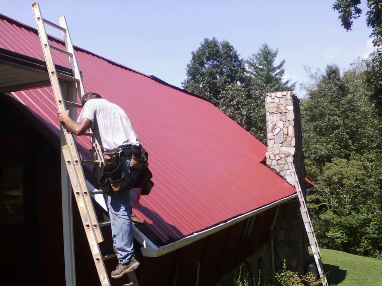 Roofer on a ladder beside a red metal roof and stone chimney — Soffit & Fascia Repair by Roofing Brothers in Holiday